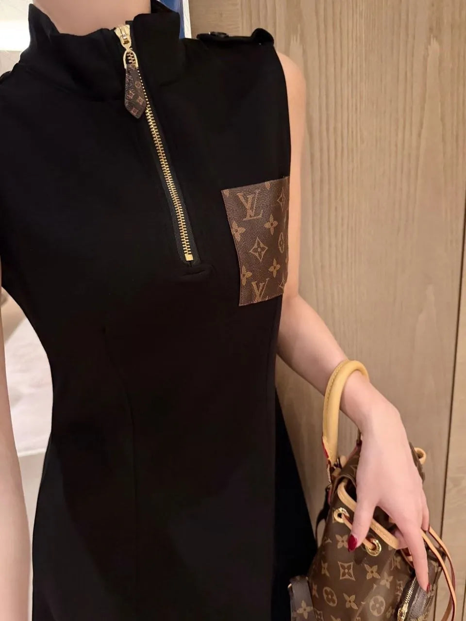 Person wearing a black sleeveless dress with a gold zipper, holding a Louis Vuitton handbag.