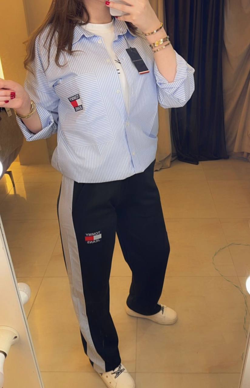 Person wearing a light blue striped shirt and dark pants with visible brand logos, standing in a room.