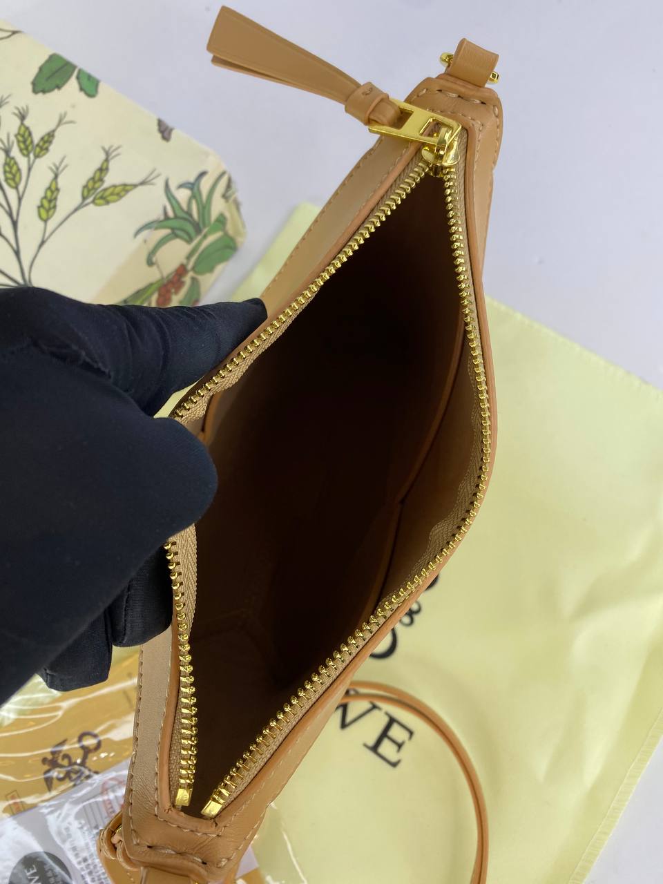 Brown leather pouch with gold zipper being held open by a black gloved hand, with a patterned background.