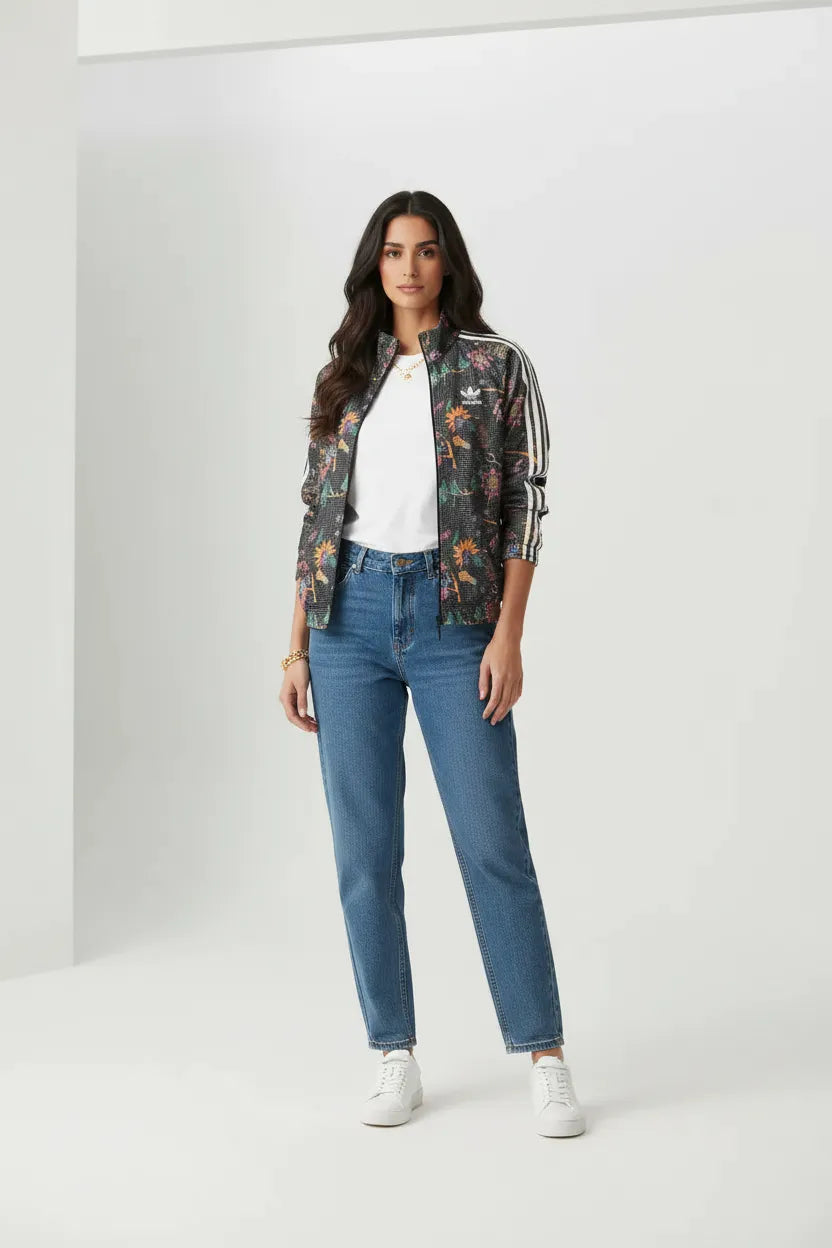 Female model wearing Adidas Originals black floral track jacket open white t-shirt blue relaxed jeans white sneakers gray studio background