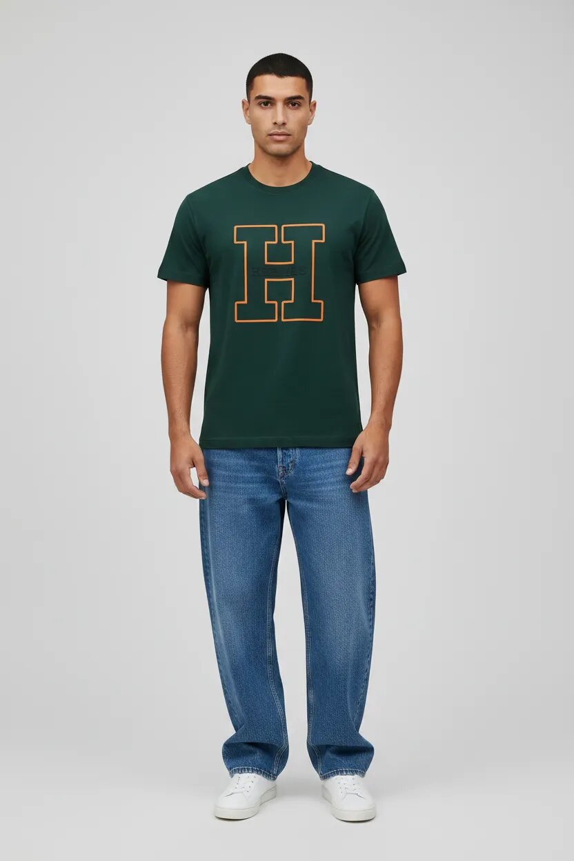 Male model wearing dark green H logo graphic t-shirt blue wide leg jeans white sneakers gray studio background men's fashion editorial