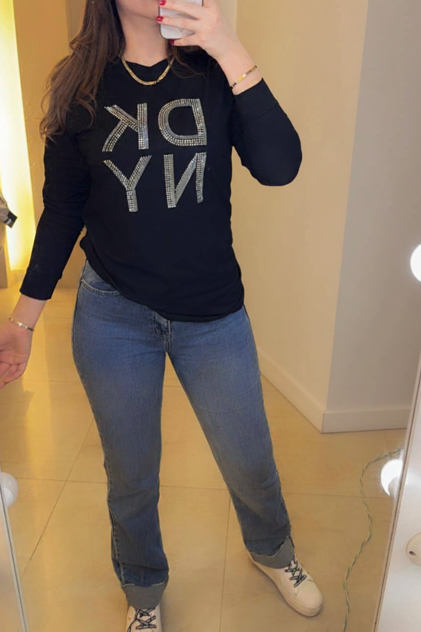 Female model wearing DKNY black rhinestone NY logo shirt blue tapered jeans white sneakers light gray studio background