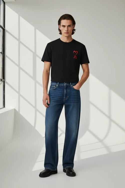 Man wearing a black t-shirt with a red design and blue jeans standing in a well-lit room.