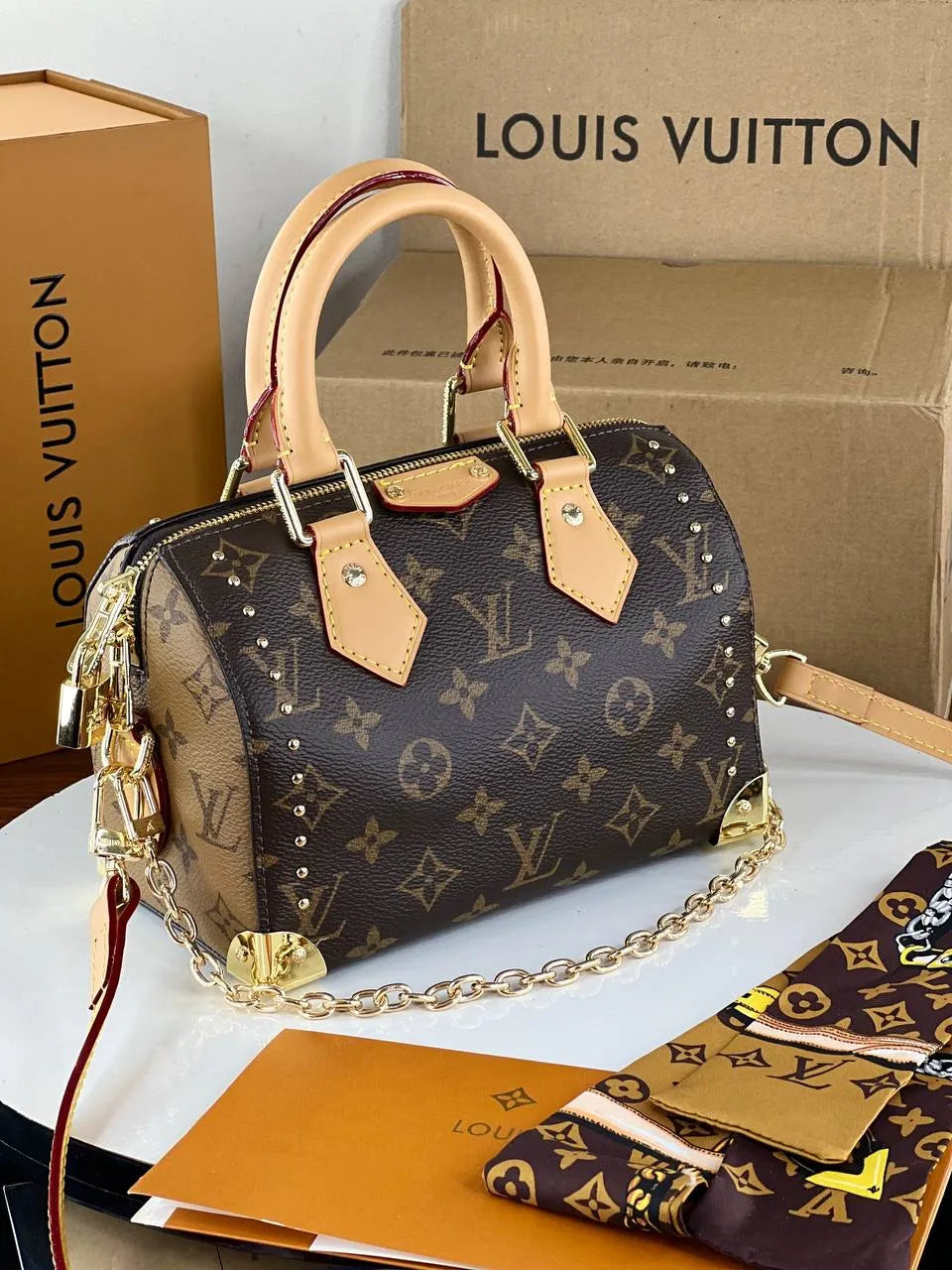 Louis Vuitton handbag with monogram pattern, beige handles, and gold chain on a white surface with branded boxes in the background.