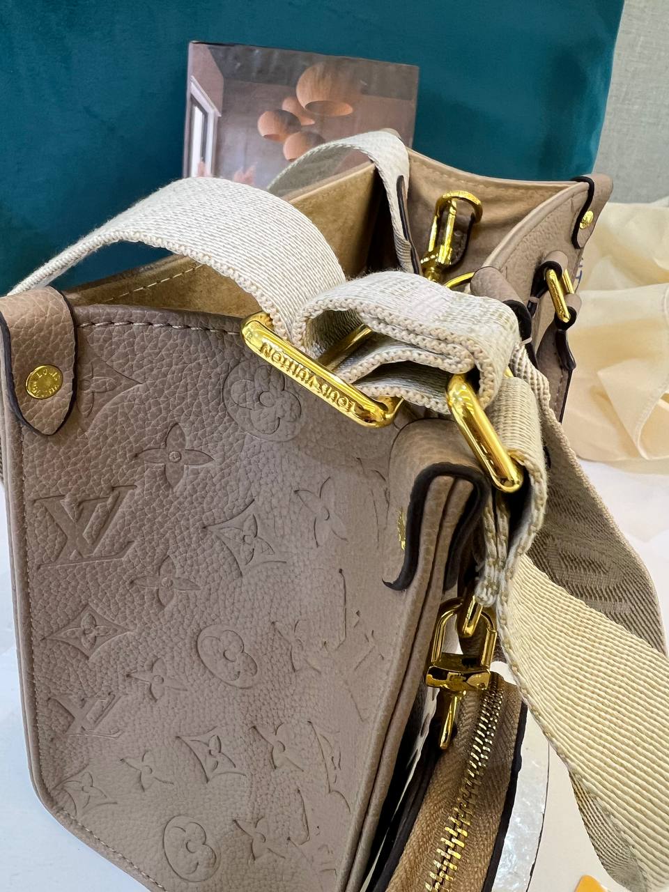 Beige handbag with gold zippers and monogram pattern, against a blurred background.