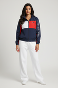 Female model wearing Tommy Hilfiger colorblock windbreaker jacket zipped closed white wide leg sweatpants white sneakers gray studio background