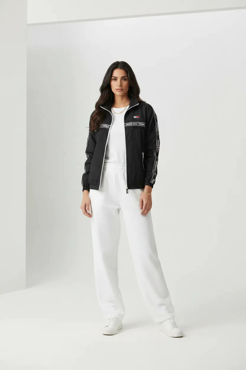 Female model wearing Tommy Jeans black windbreaker jacket open white t-shirt white wide leg sweatpants white sneakers gray studio background
